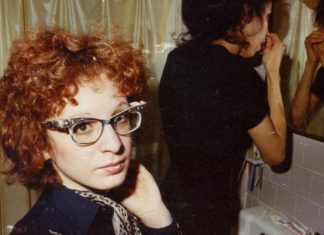 Golden Lion for documentary about Nan Goldin | Movies | DW A woman with curly red hair and glasses is standing in the bathroom; another woman is standing at the mirror in the background and doing something in her face.