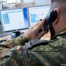 A uniformed Bundeswehr soldier makes a phone call while sitting at a desk with two computer screens.