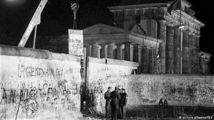 Opening of the Wall at the Brandenburg Gate: Police officers from East and West Berlin talk at the Brandenburg Gate while a piece of the Wall is being lifted up behind them with a crane