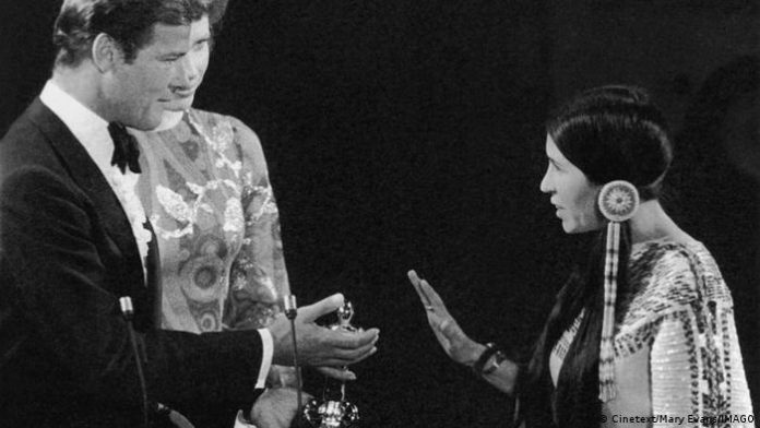 Sacheen Littlefeather makes a defensive hand gesture, Roger Moore looks at her with trophy in hand, Liv Ullman in the middle